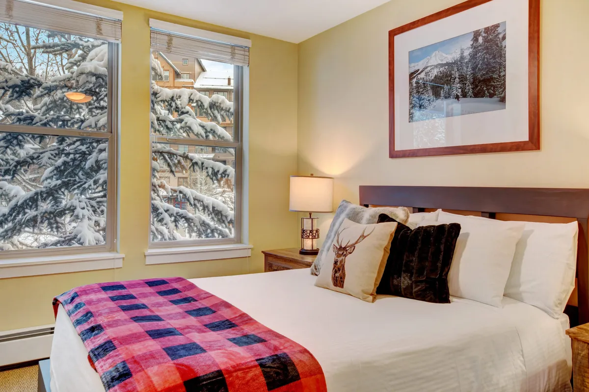 A bedroom featuring a bed draped with a plaid blanket, a nightstand with a lamp, framed mountain photography on the wall, and windows that reveal a snowy, scenic view. The space feels warm and inviting, perfect for unwinding after a day outdoors.