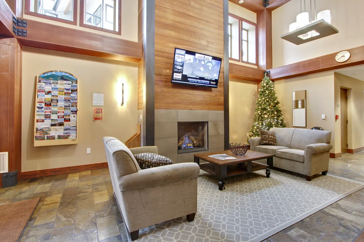 A spacious, warmly furnished resort lobby featuring a prominent fireplace, comfortable seating area, decorative tree, and television. The room exudes a welcoming, social atmosphere, inviting guests to relax, gather, or enjoy a quiet moment by the fire.
