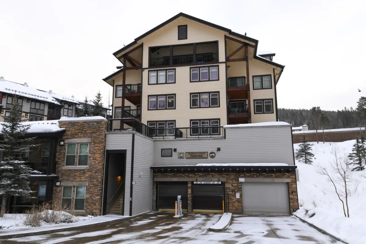 The image displays the resort's main entrance, featuring expansive windows and notable roof architecture. Snow is visible, suggesting a winter setting, while the cozy ambiance and welcoming design evoke the feel of arriving at a high-end mountain lodge. There are hints of social activity, as this is a gathering point after outdoor adventures.