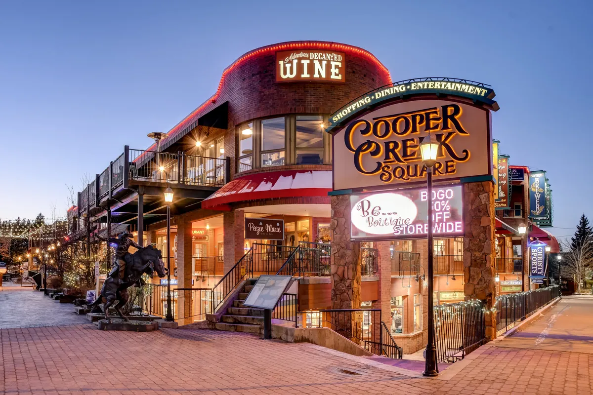 A lively exterior scene showing Cooper Creek Square, with clear signage, outdoor dining spaces, and a distinctive horse sculpture. The area appears vibrant and social, with opportunities for shopping, dining, and community events.