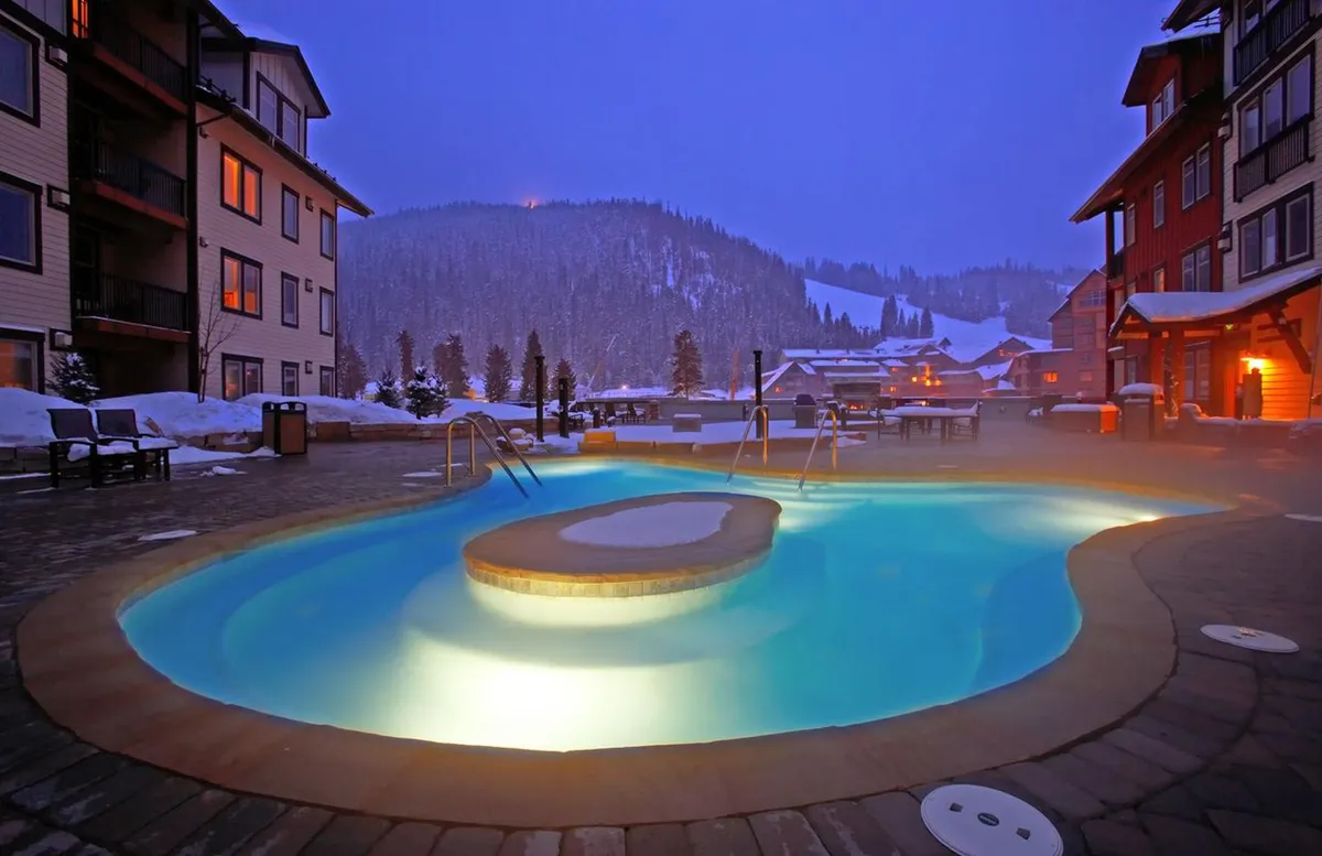 A large outdoor hot tub set amidst a snowy mountain landscape, surrounded by warmly lit resort buildings. Several guests are relaxing in the steaming water, while others chat and enjoy the winter evening. The inviting ambiance is enhanced by the contrast of the steamy water against the crisp outdoor air.