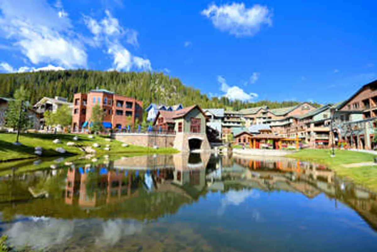 This image captures a picturesque exterior scene with reflections in a tranquil pond, surrounded by charming buildings, lush green landscape, and a bright blue sky dotted with clouds. The scene invites viewers to imagine leisurely strolls along the water, outdoor activities, or simply relaxing while taking in the mountain ambiance.