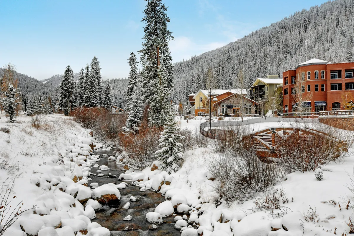 A tranquil exterior shot capturing snow-covered trees along a stream, colorful buildings, and a wooden bridge. The scene suggests easy access to nature and the picturesque surroundings of the resort and base village in wintertime.