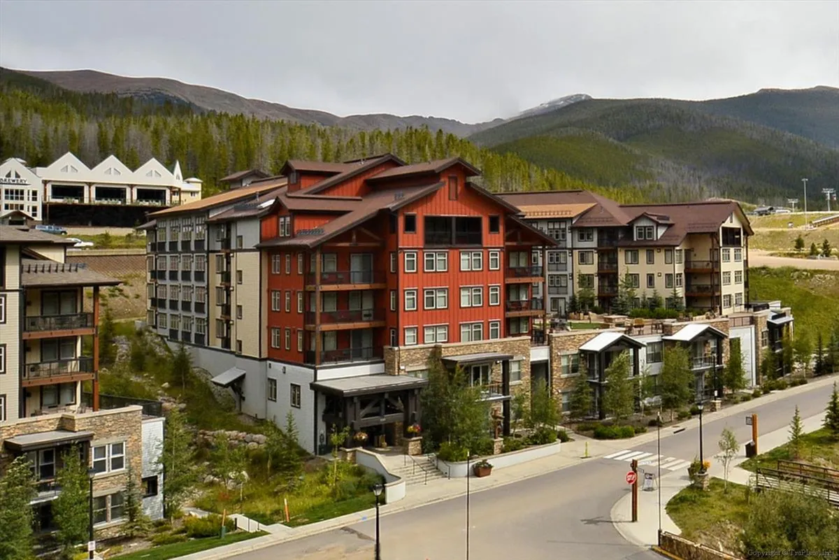 An inviting exterior view of a tall red and beige building surrounded by lush greenery, with majestic mountains as the backdrop. The scene includes a quiet street lined with light poles, and hints of guests walking or heading toward nearby trails.
