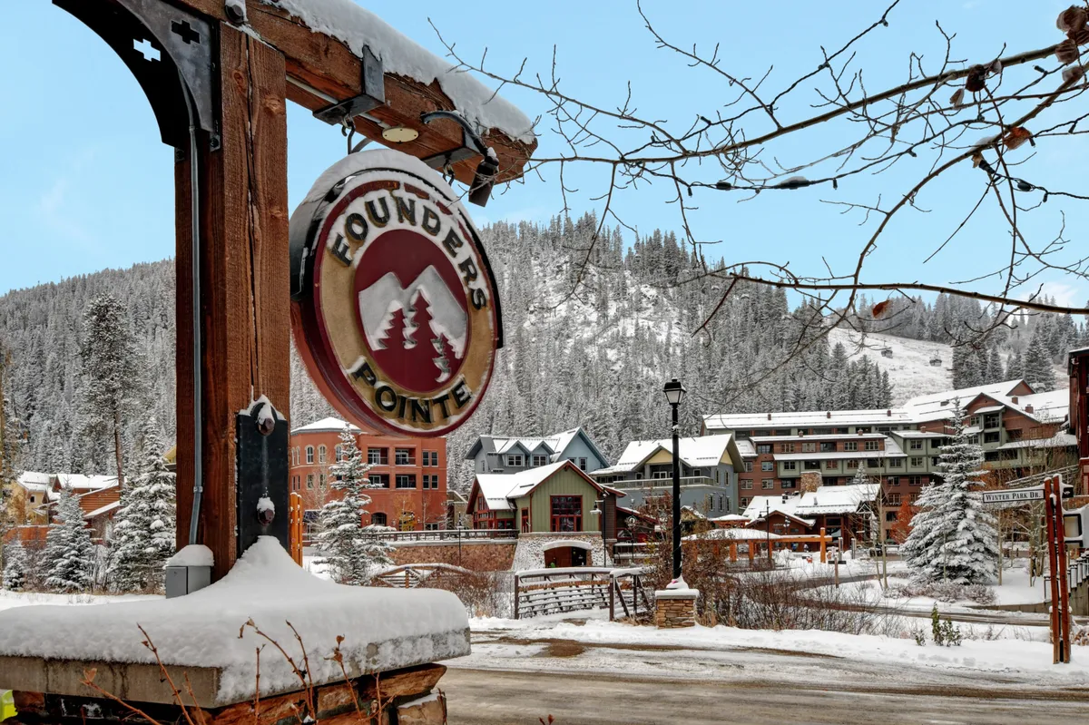 An inviting exterior view of the base village with snow-covered trees, colorful buildings, and a welcoming sign. The scene captures the spirit of a ski resort community in winter, with hints of activity and togetherness in a picturesque mountain setting.