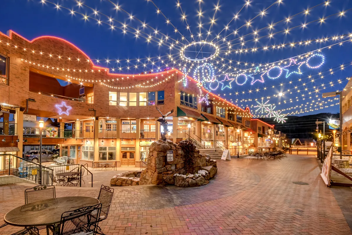 A lively exterior scene of a resort village at dusk, adorned with decorative lights, welcoming storefronts, and a central fountain where people gather and socialize. The festive ambiance hints at the vibrancy and community spirit of the base village during peak seasons.