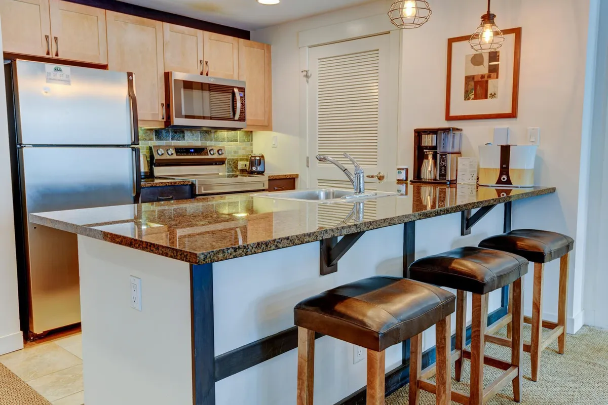 A spacious, modern kitchen featuring granite countertops, stylish bar stools, and a full range of kitchen appliances. Warm artwork adorns the wall, and the layout encourages interaction across the countertop, ideal for shared meal preparation and casual conversations.