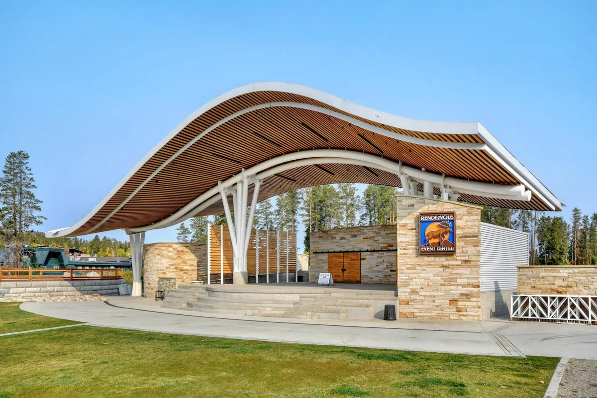 An exterior shot featuring a distinctive wave-shaped roof structure set against stone walls and wooden accents. The open-air venue is ideal for gatherings, concerts, and social community events, with a lively, festive atmosphere that suggests shared outdoor experiences.