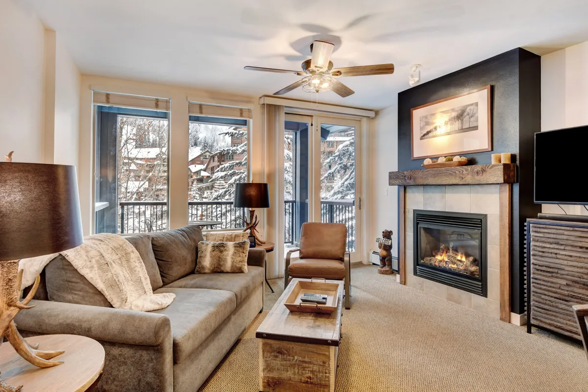 The image showcases a cozy living space featuring a crackling fireplace, a plush sofa, and expansive windows framing views of falling snow. The inviting setting is perfect for relaxing with a warm drink, reading, or socializing after a day of mountain adventures.