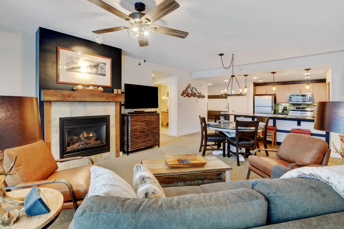 This image captures the heart of the living space featuring a rustic fireplace, comfortable sofas, a dining table, and a glimpse of the kitchen area. The setting is warm and inviting, with the fire as a focal point for cozy gatherings, while the open layout encourages connection among guests.