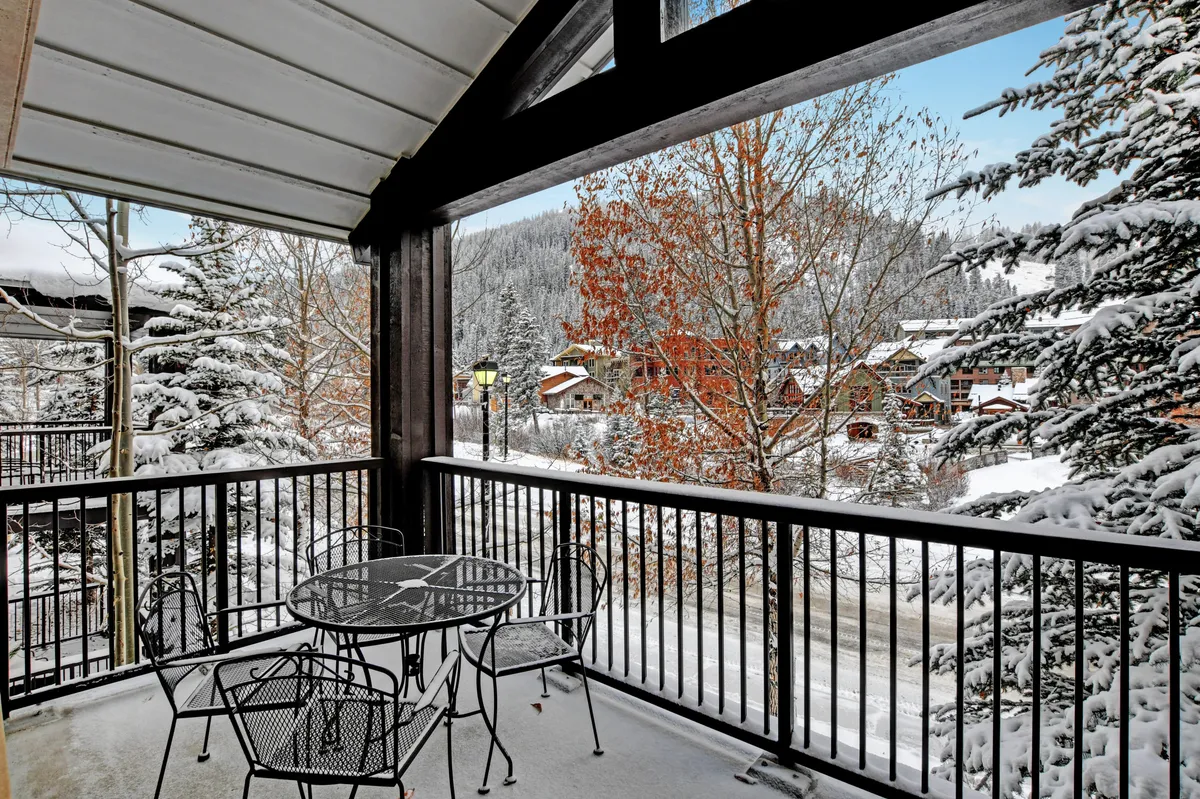 A private covered deck with a metal patio table and black chairs overlooks a wintry, snow-covered landscape and village buildings. The scene evokes peaceful moments spent sipping coffee or watching the bustling village below.